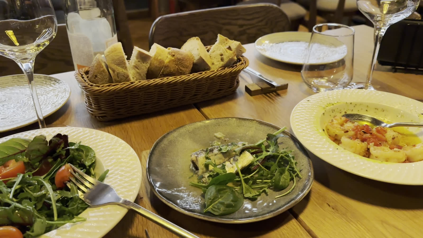 Farfurii pe masa de lemn: salate, pâine în coș, pahare de vin